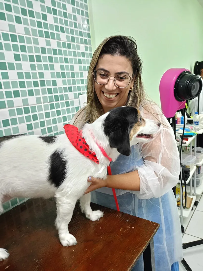 Sara sorrindo com um cachorrinho branco e preto de bandana vermelha na mesa de tosa